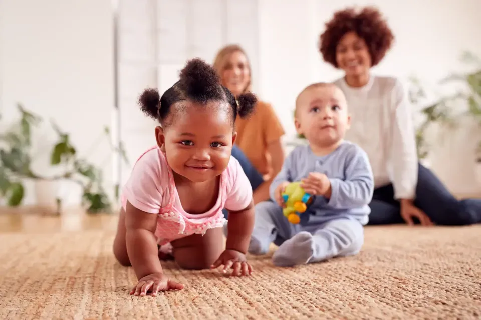 Smiling toddler crawling on a carpet with another baby nearby and two adults supervising in the background during a playdate.
